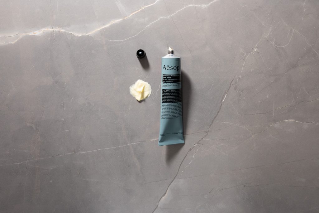 Aesop Reverence Aromatique Hand Balm with cream swatch on grey stone background.