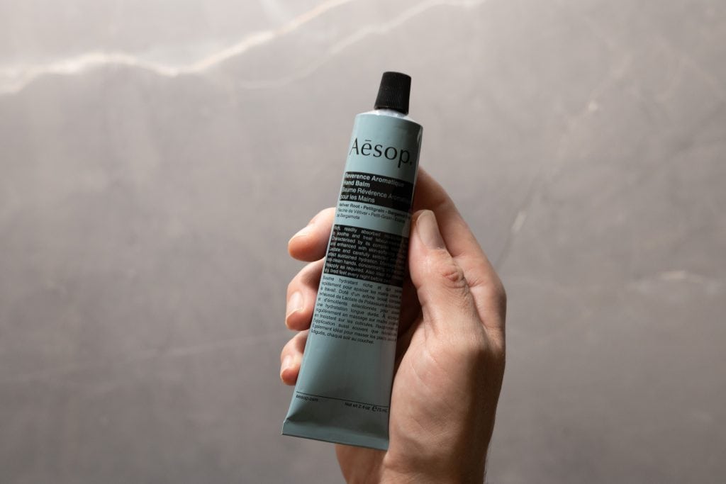 Hand holding Aesop Reverence Aromatique Hand Balm against grey background.
