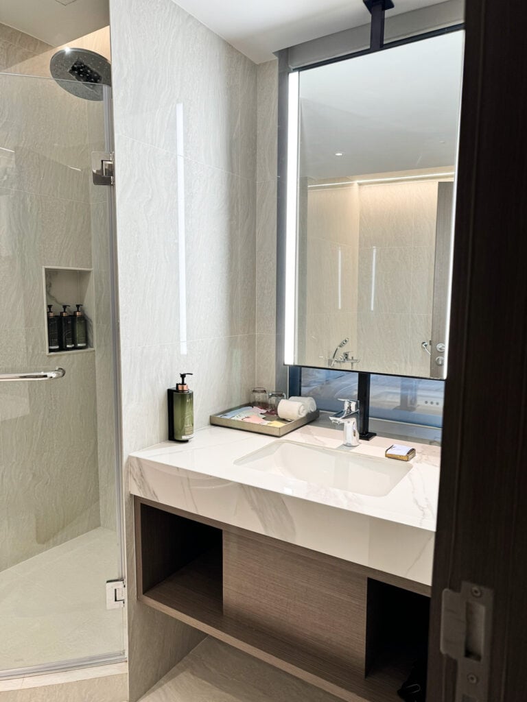 Bathroom vanity at Pathumwan Princess Hotel with sink, backlit mirror, and fixed shower products visible.