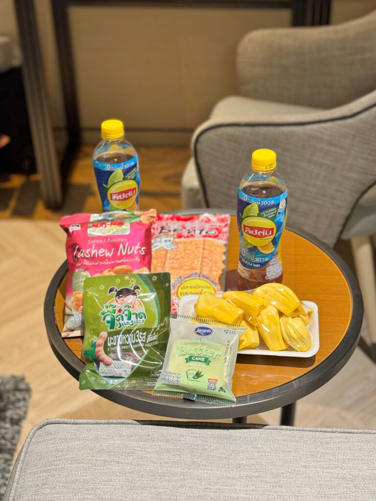 In-room snack table at Pathumwan Princess Hotel with iced-tea, cashew nuts, tamarind sweets, pandan cake, and fruit on a wooden bench