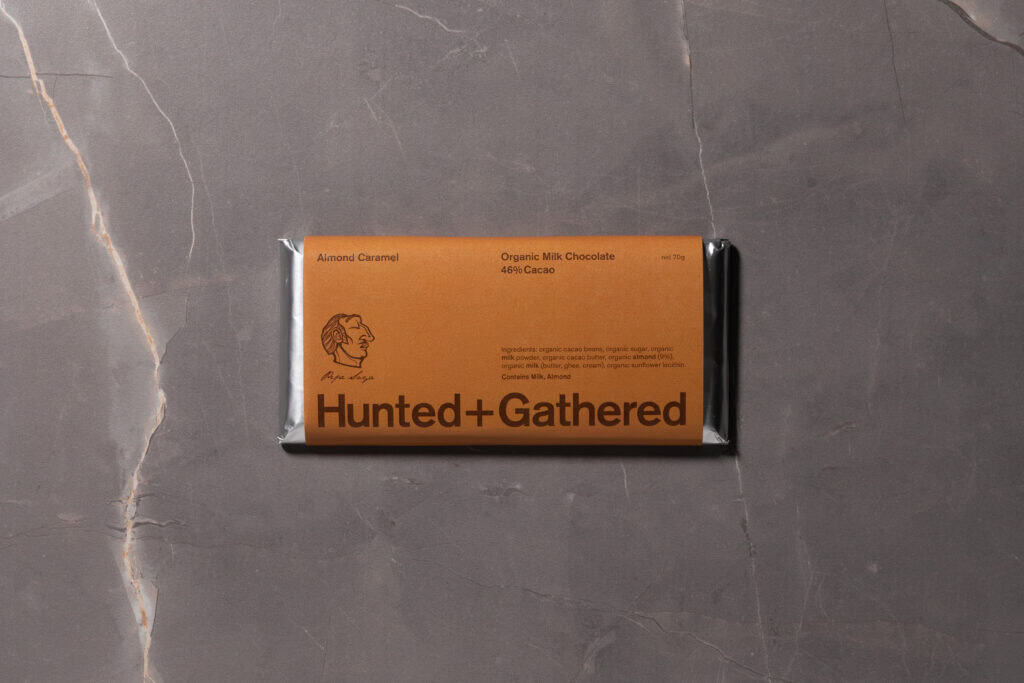 Hunted + Gathered Almond Caramel 46% Cacao Milk Chocolate bar in minimalist brown packaging on grey marble background.