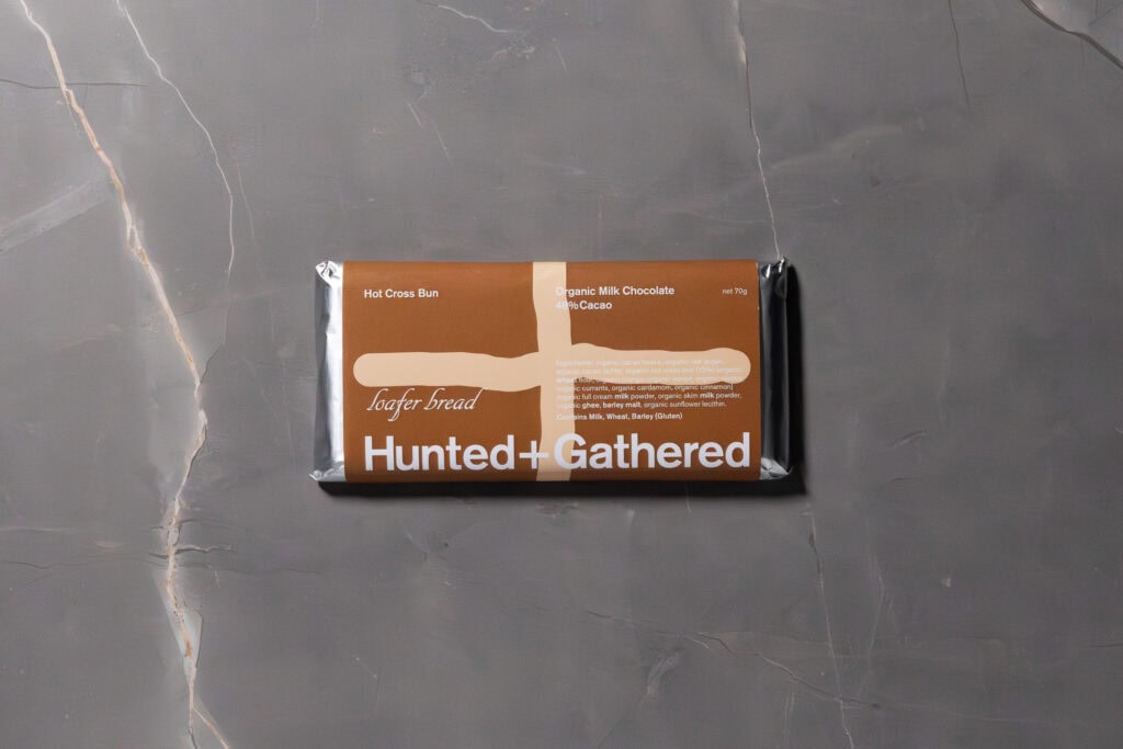 Hunted + Gathered Milk Chocolate + Hot Cross Bun bar in packaging, placed on a grey stone surface.
