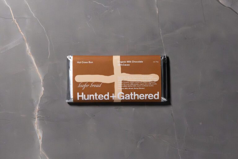 Hunted + Gathered Milk Chocolate + Hot Cross Bun bar in packaging, placed on a grey stone surface.