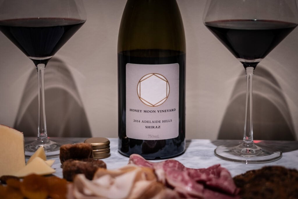 Bottle of Honeymoon Vineyard Shiraz 2014 flanked by two red wine glasses, with figs, charcuterie, and cheese in soft foreground.