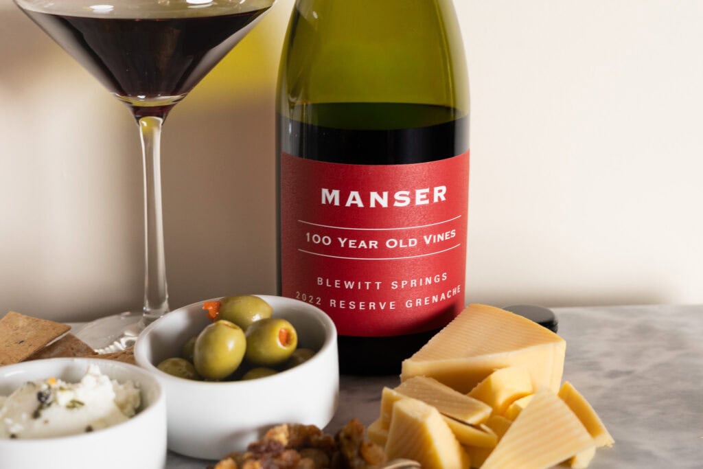 Manser 100 Year Old Vines Reserve Grenache 2022 bottle with wine glass, cheese, olives, and crackers