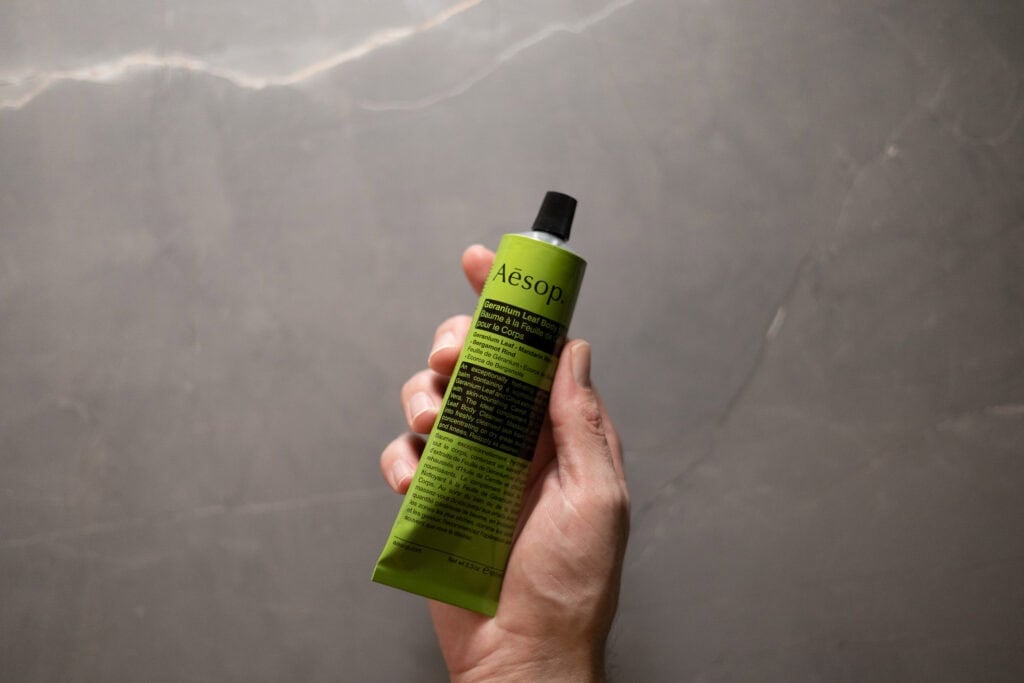 Hand holding a tube of Aesop Geranium Leaf Body Balm against a grey stone background.