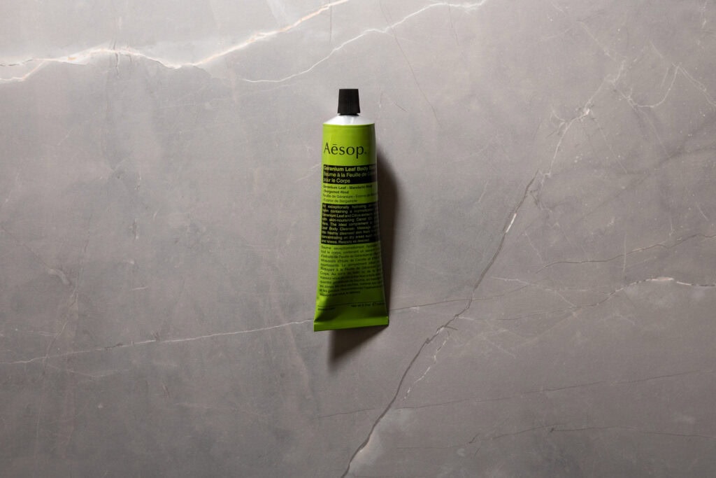Green tube of Aesop Geranium Leaf Body Balm on a grey stone surface.
