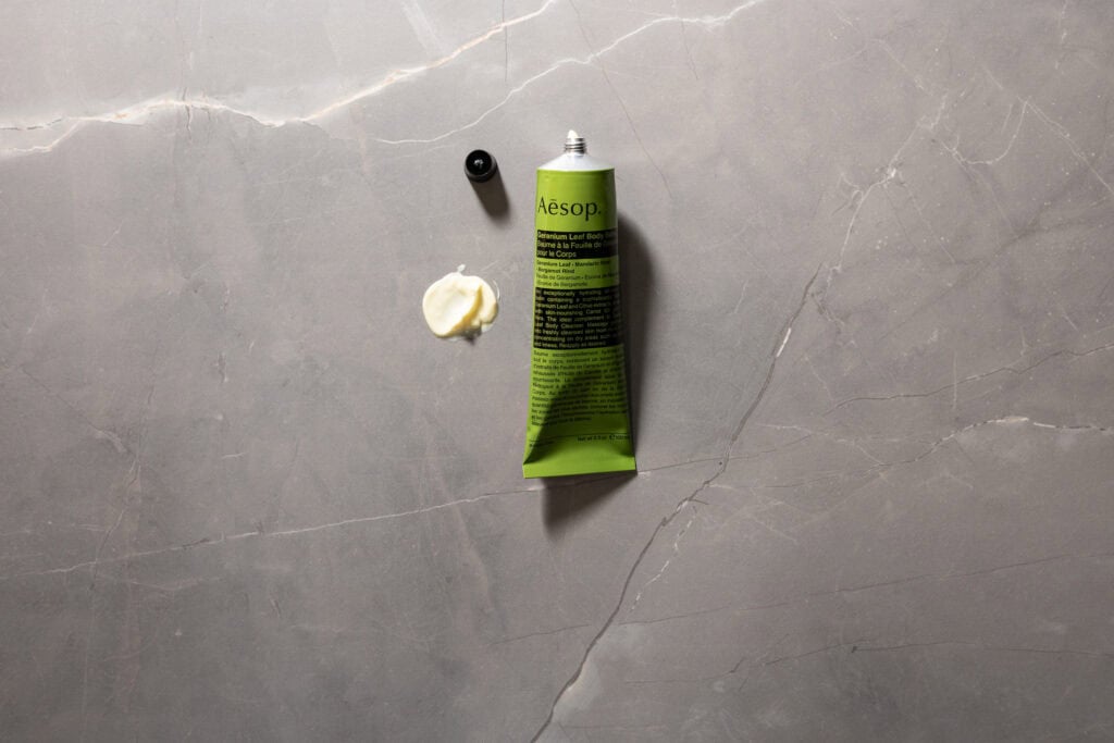 Aesop Geranium Leaf Body Balm with the cap off and a small amount of balm displayed on a grey stone surface.