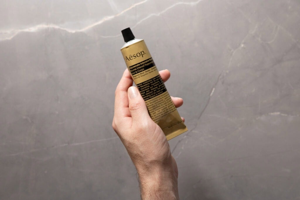 Hand holding a tube of Aesop Rejuvenate Intensive Body Balm against a grey stone background.