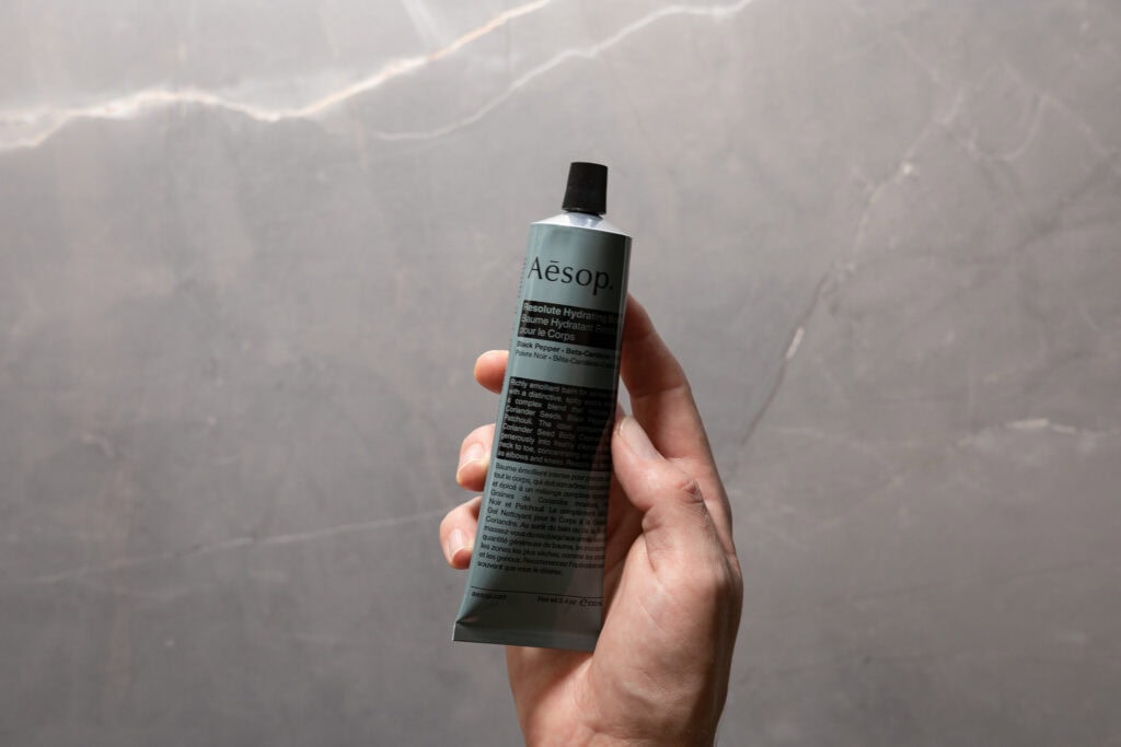 Hand holding a tube of Aesop Resolute Hydrating Body Balm against a grey stone background