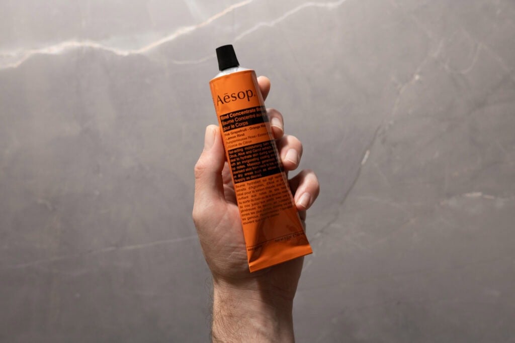 Hand holding an Aesop Rind Concentrate Body Balm tube against a grey stone background.