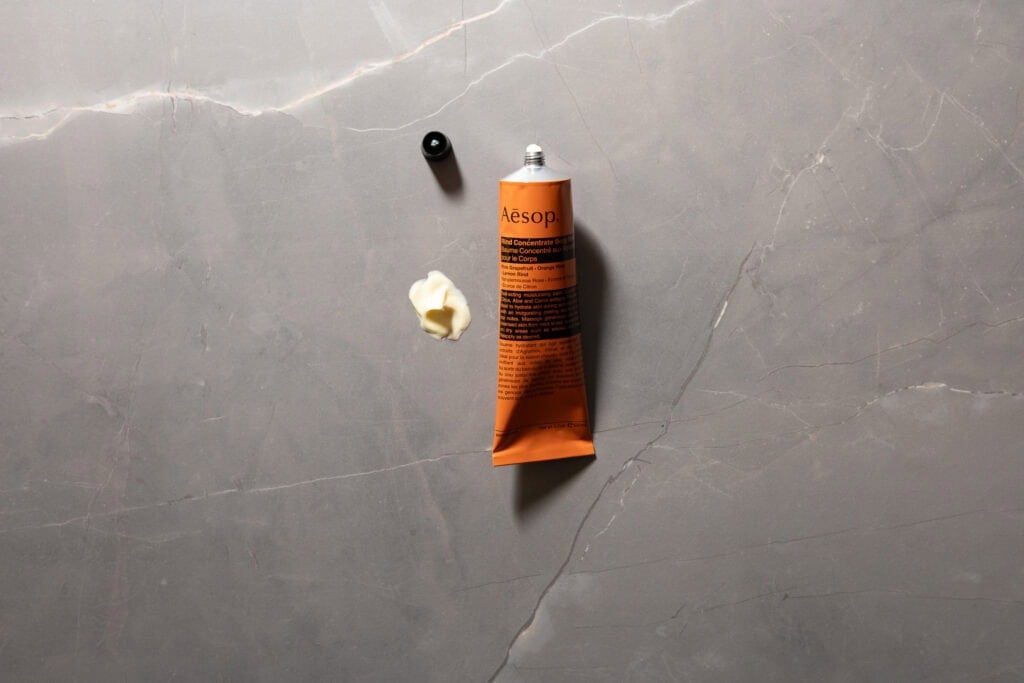 Aesop Rind Concentrate Body Balm with the cap off and a small amount of balm displayed on a grey stone surface.