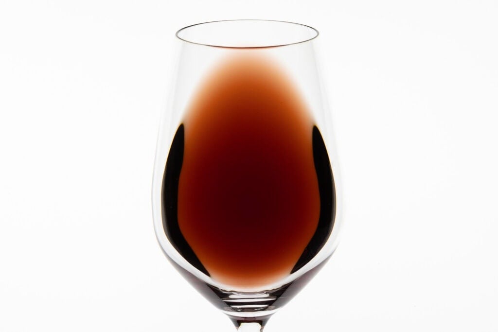 A close-up of Bannockburn Geelong Pinot Noir 2024 in a clear wine glass against a bright white background, showing its deep red core fading to a lighter, translucent red towards the edges.