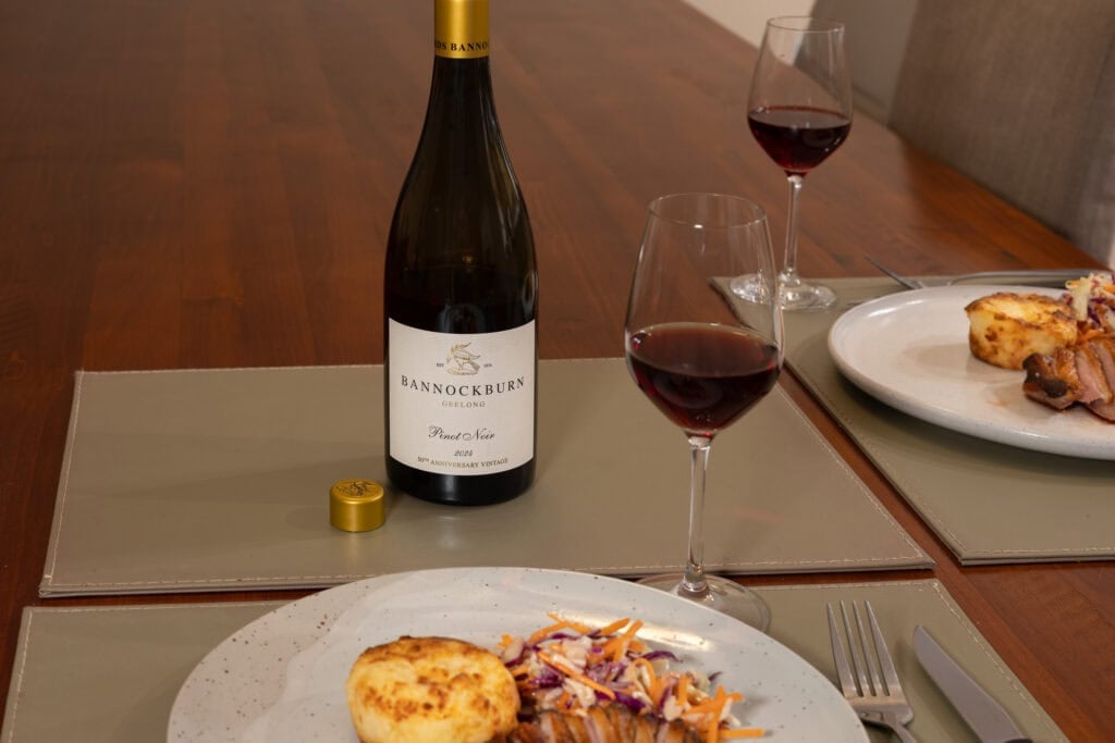 A bottle and two glasses of Bannockburn Geelong Pinot Noir 2024 on a wooden dining table, served alongside plates of pan-fried duck breast, potato gratin and coleslaw.
