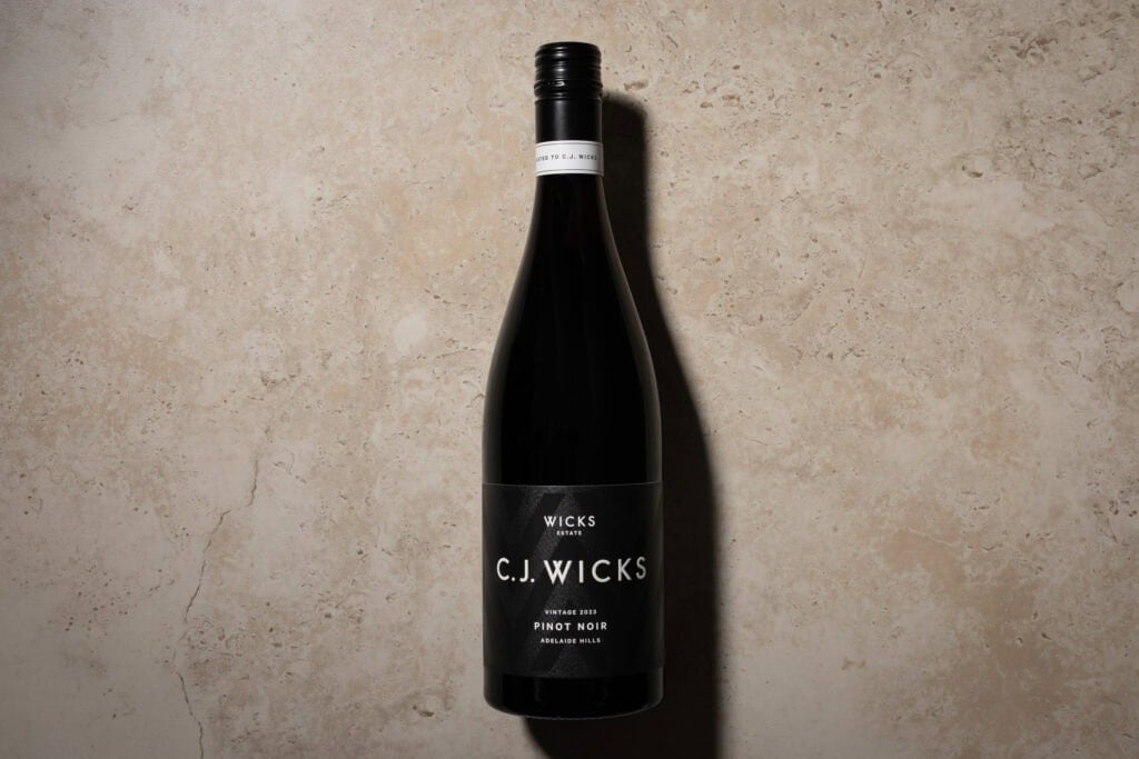 Wicks Estate C.J. Wicks Pinot Noir 2023 bottle standing against a pale stone background, with a black label and long shadow.