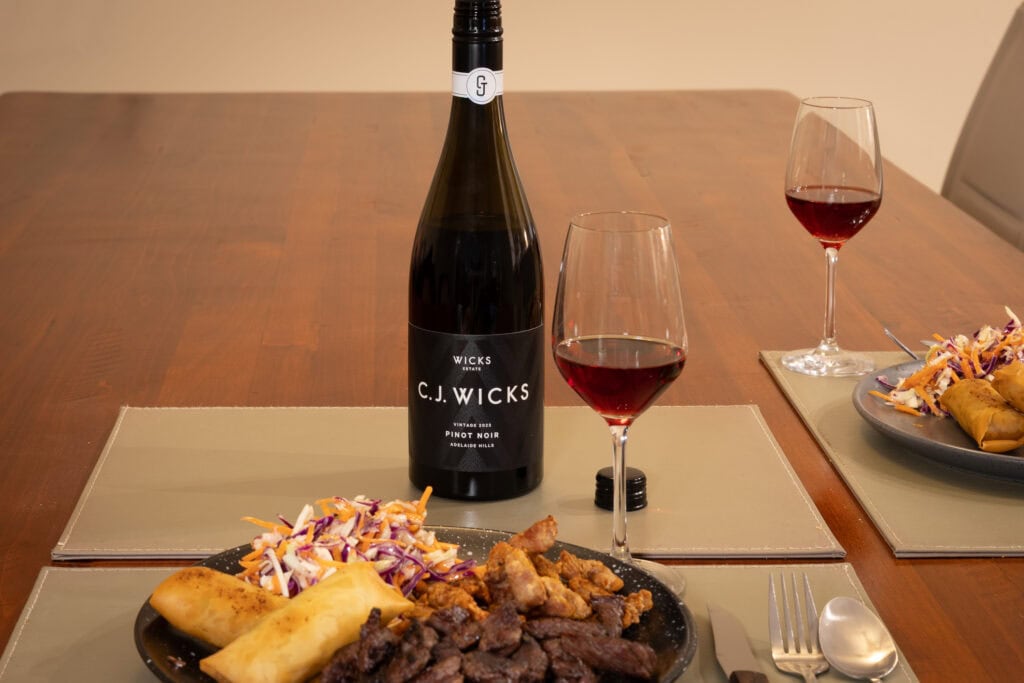 Wicks Estate C.J. Wicks Pinot Noir 2023 served with rump beef, tempura pork loin, spring rolls and coleslaw at a dining table.