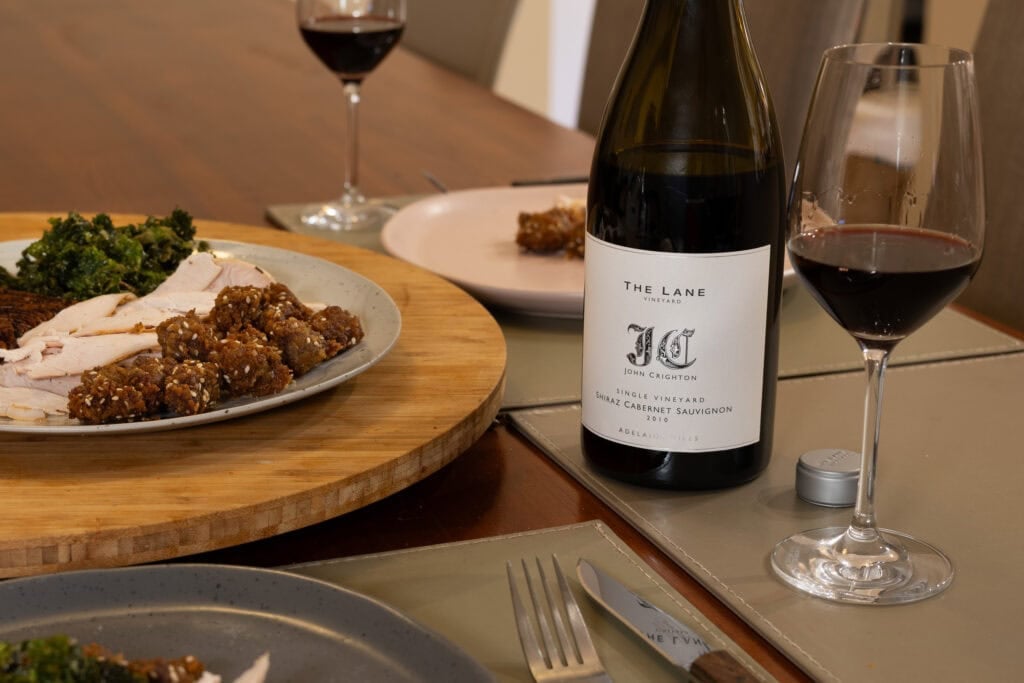 Bottle and glass of The Lane John Crighton 2010 beside plates of roast chicken, fried kale and meat dishes