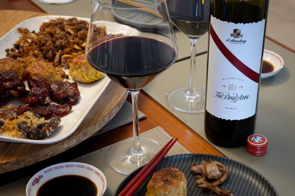 Bottle of d’Arenberg The Dead Arm Shiraz 2019 beside wine glasses and fried savoury dinner dishes